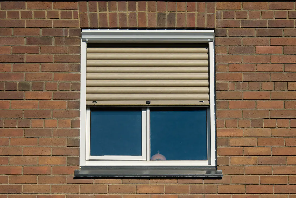 Inbouwrolluik boven een nieuw wit kozijn op een bakstenen gevel, half neergelaten