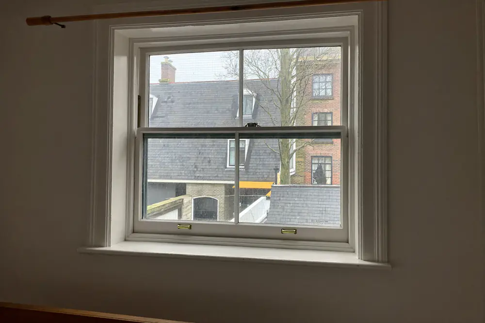 Wit houten schuifraam met kleine ruiten en koperen sluitingen, van binnen gezien