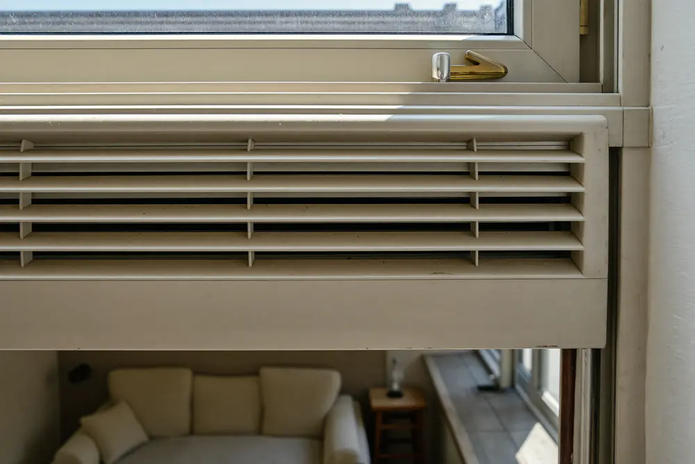 Klein ventilatierooster in de bovenkant van een wit kozijn, van binnen gezien